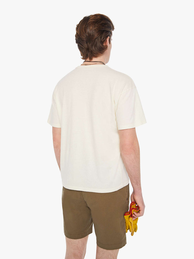 Back view of a man in a cream oversized tee with drop shoulders and a loose fit that features a faded dog graphic with colorful text on the front.