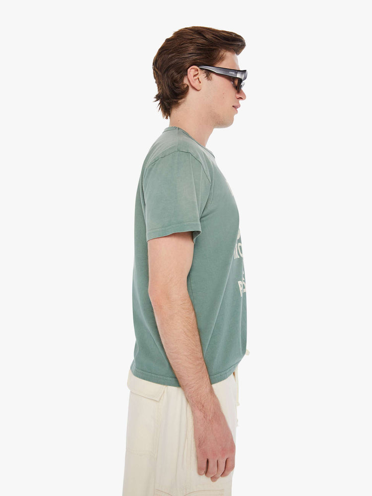 Side view of a man in a pale green oversized tee with drop shoulders and a loose fit featuring an off-white faded text graphic.
