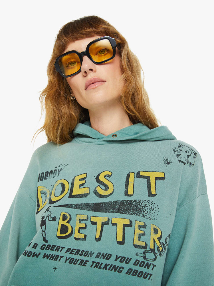 WOMEN close up view of an teal oversized hoodie with drop shoulders, ribbed hems and a front patch pocket and colorful text, doodles, and fading at the shoulders.