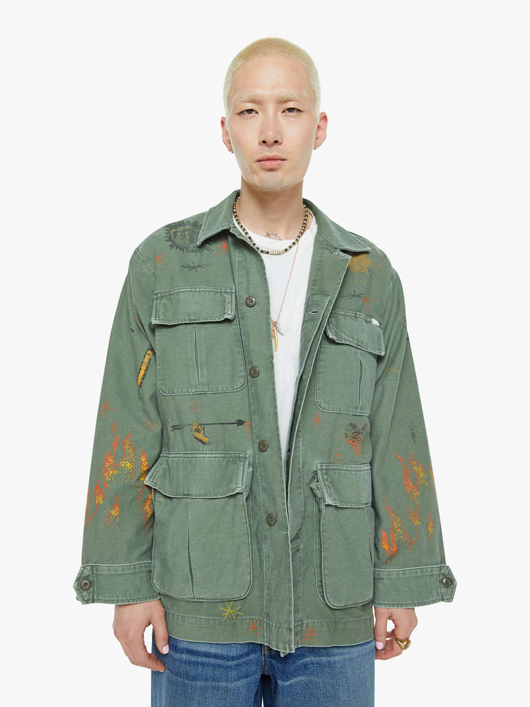 Front view of a men in a faded army-green hue military-inspired jacket with a collar, front patch pockets with painterly screen-printed details throughout.