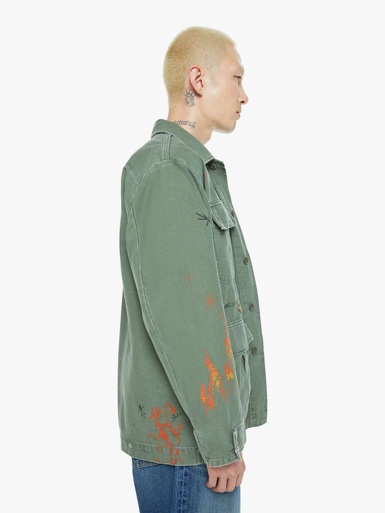 Size view of a men in a faded army-green hue military-inspired jacket with a collar, front patch pockets with painterly screen-printed details throughout.