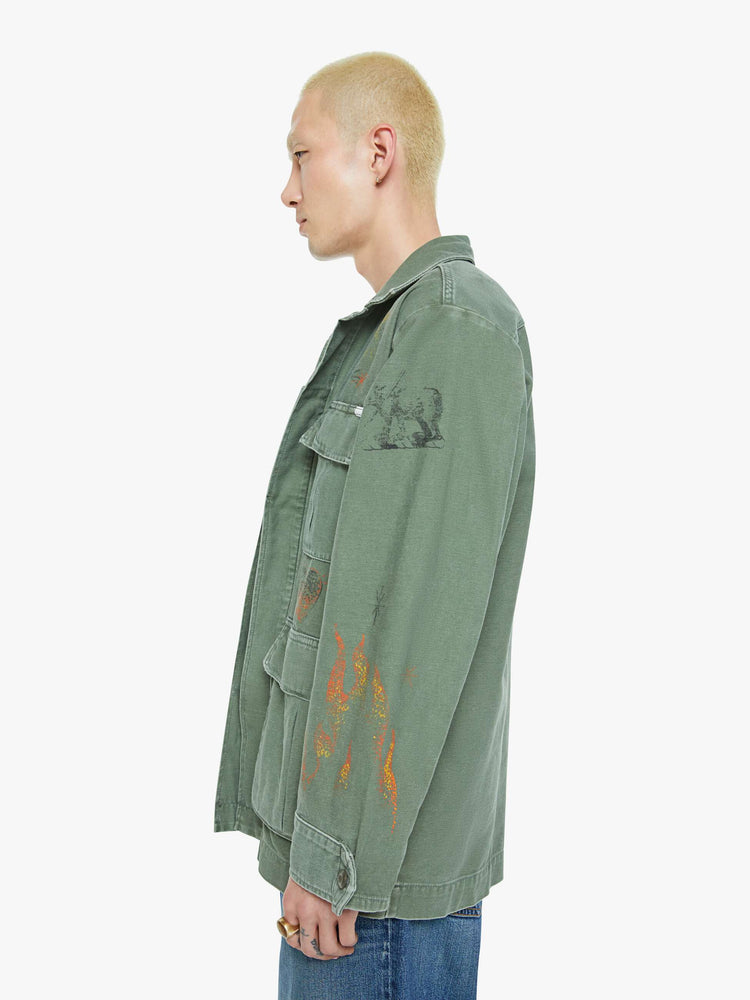 Right side view of a men in a faded army-green hue military-inspired jacket with a collar, front patch pockets with painterly screen-printed details throughout.