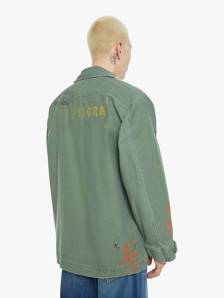 Back view of a men in a faded army-green hue military-inspired jacket with a collar, front patch pockets with painterly screen-printed details throughout.