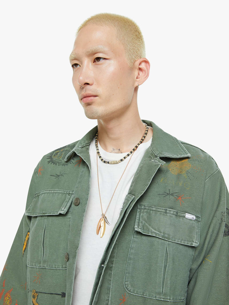 Close up view of a men in a faded army-green hue military-inspired jacket with a collar, front patch pockets with painterly screen-printed details throughout.