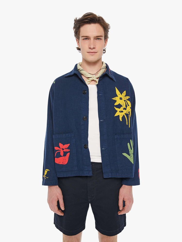 Front view of a man in a dark navy vintage-inspired chore jacket with slightly dropped shoulders, oversized patch pockets and a boxy fit with colorful floral graphics throughout.