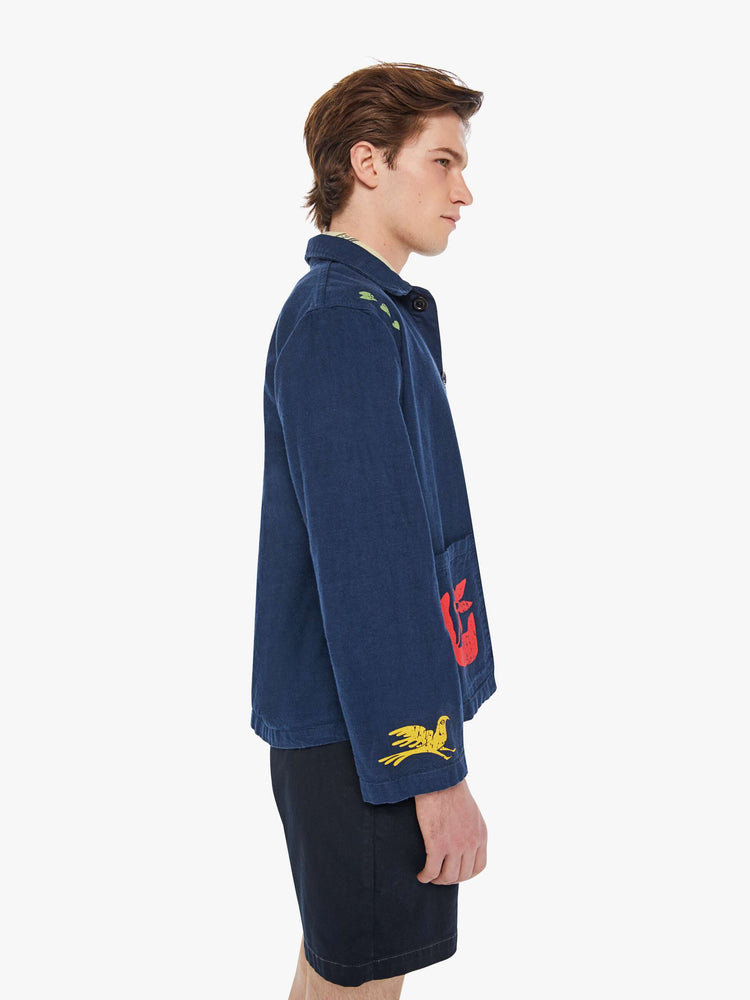 Right side view of a man in a dark navy vintage-inspired chore jacket with slightly dropped shoulders, oversized patch pockets and a boxy fit with colorful floral graphics throughout.