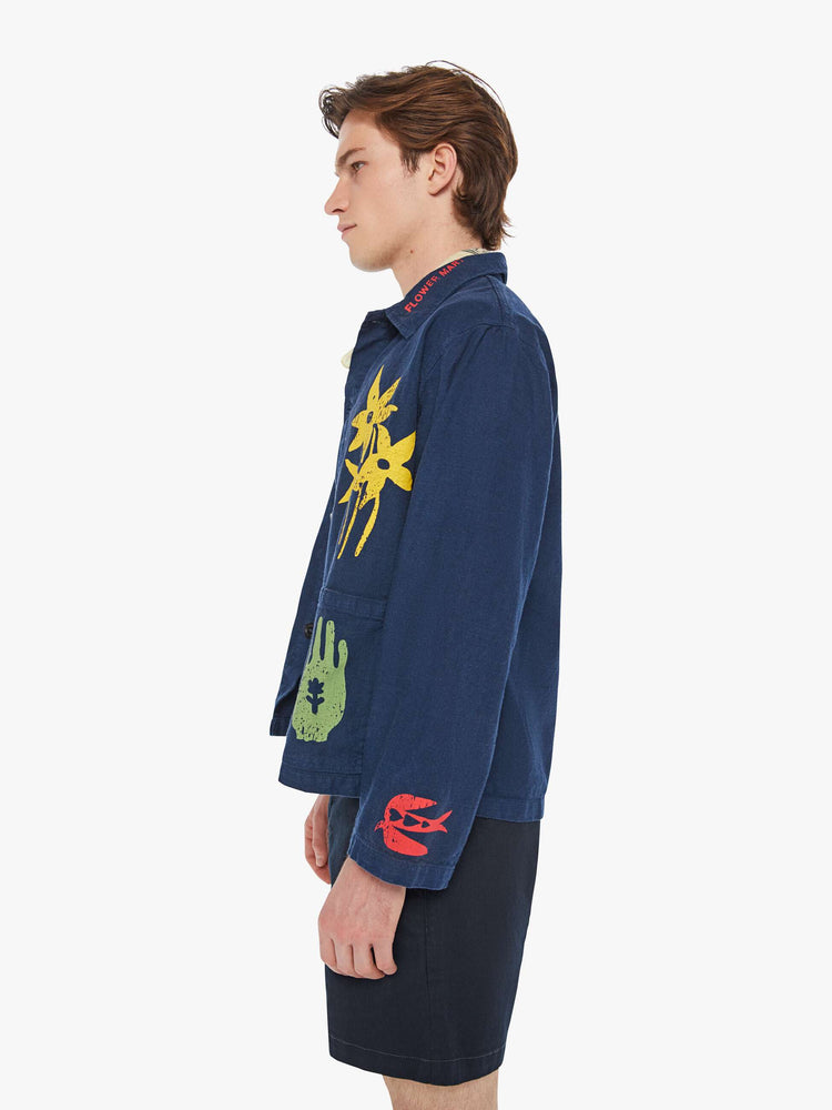 Left side view of a man in a dark navy vintage-inspired chore jacket with slightly dropped shoulders, oversized patch pockets and a boxy fit with colorful floral graphics throughout.