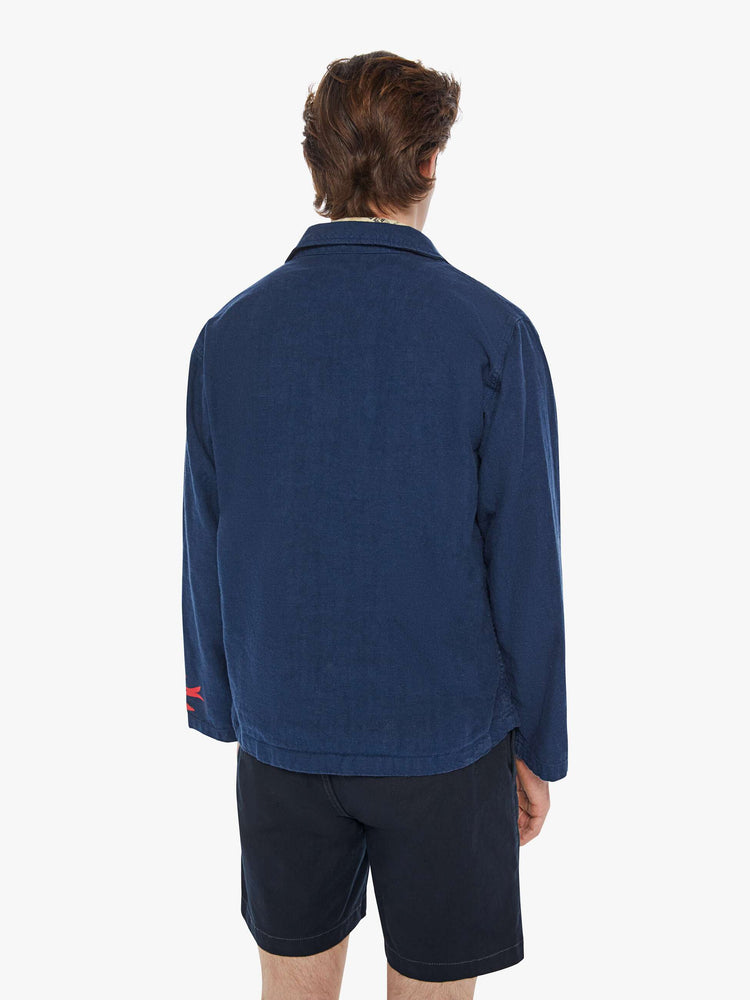 Back view of a man in a dark navy vintage-inspired chore jacket with slightly dropped shoulders, oversized patch pockets and a boxy fit with colorful floral graphics throughout.