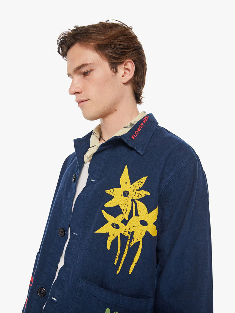 Detailed view of a man in a dark navy vintage-inspired chore jacket with slightly dropped shoulders, oversized patch pockets and a boxy fit with colorful floral graphics throughout.