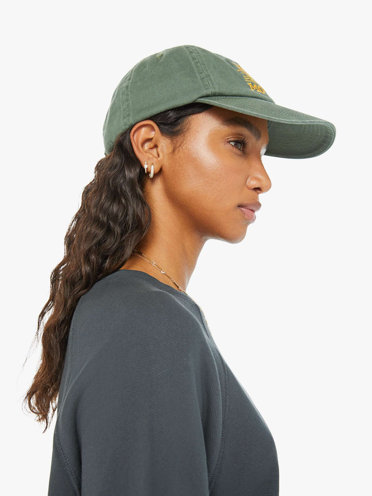 Side view of a woman in a faded sage green vintage-inspired baseball hat with a golden-yellow sunset graphic and MOTHER's name embroidered on the front.