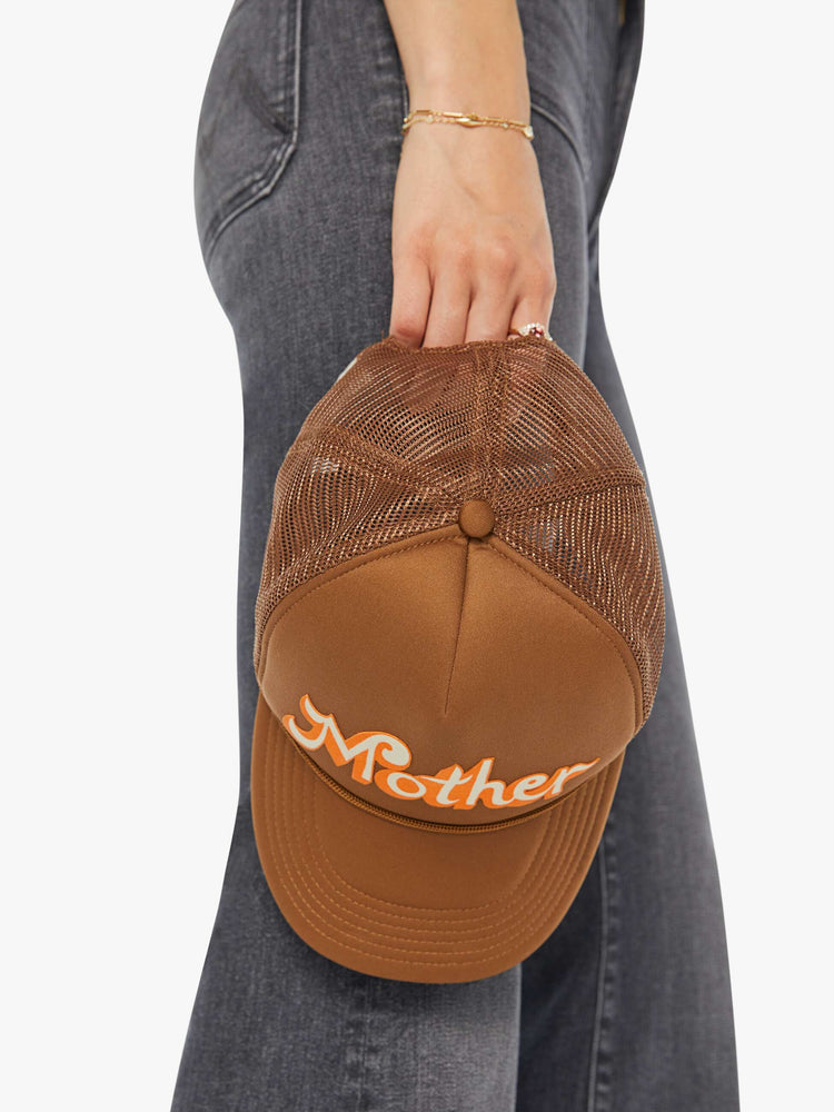 Holding view of a woman in a brown trucker hat with MOTHER's name in orange and cream on the front.