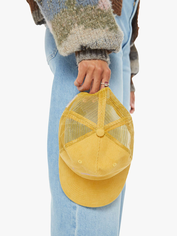 Lay down view of a model holding  a vintage-inspired trucker hat designed in mustard-yellow corduroy with MOTHER's name in lavender on the front.