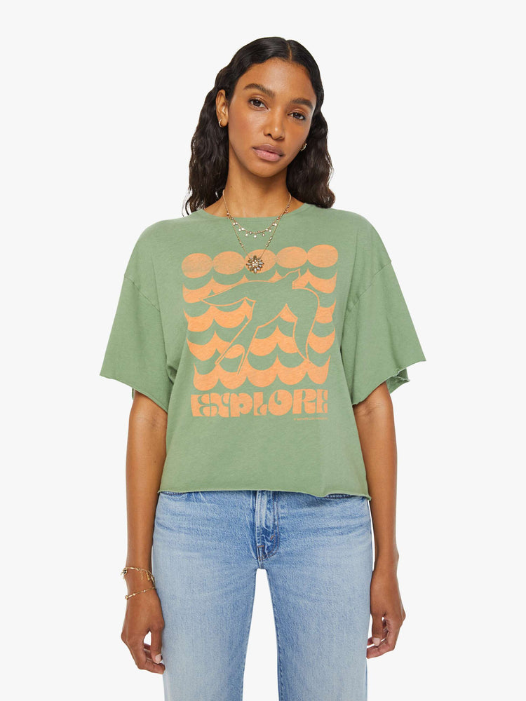 Front view of a woman in a sage green  oversized crewneck tee with drop shoulders and raw hems with a peach bird graphic with text on the front.