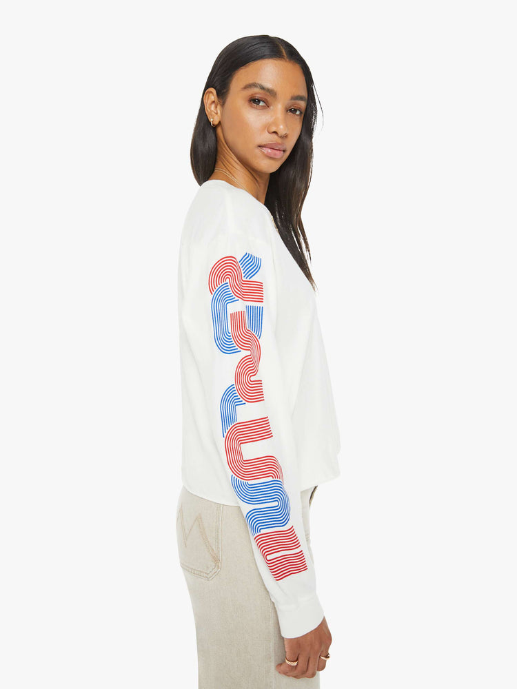 Side view of a woman in a white with red and blue graphic text down the arms and on chest oversized long sleeve crewneck with a raw cropped hem.