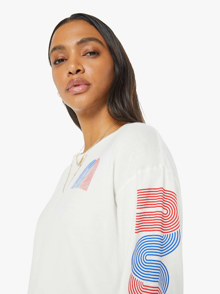 Close up view of a woman in a white with red and blue graphic text down the arms and on chest oversized long sleeve crewneck with a raw cropped hem.