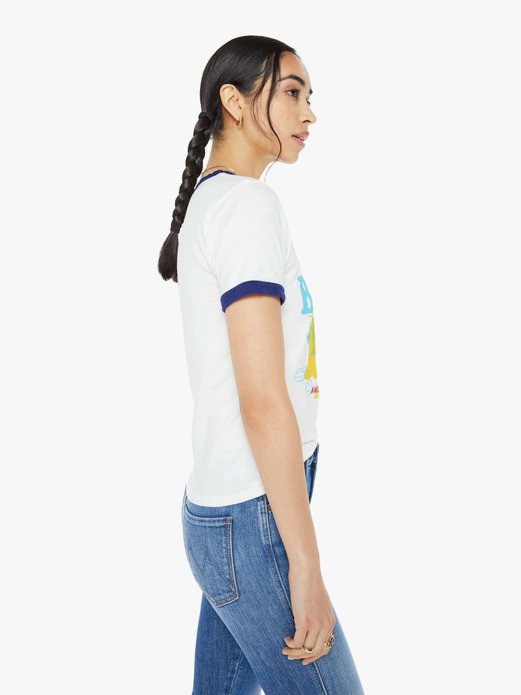 Side view of a woman in a white tee features a text graphic in yellow, blue and red on the front and navy trim on the neck and sleeves.