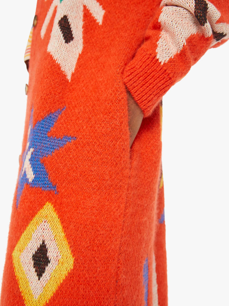 Detailed view of a woman in a long bright orange duster cardigan with colorful motifs, a crew neck, drop shoulders and buttons down the front.