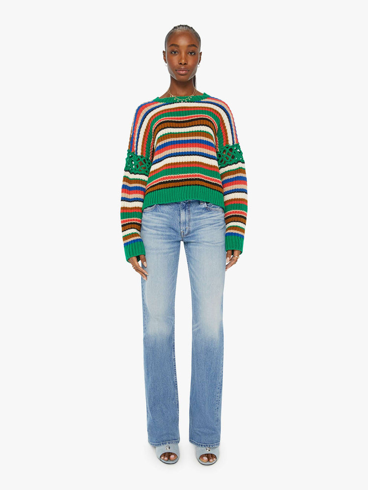 Full front view of a woman in a slightly cropped crewneck sweater in a colorful horizontal stripe pattern with green crochet panels on the sleeves.