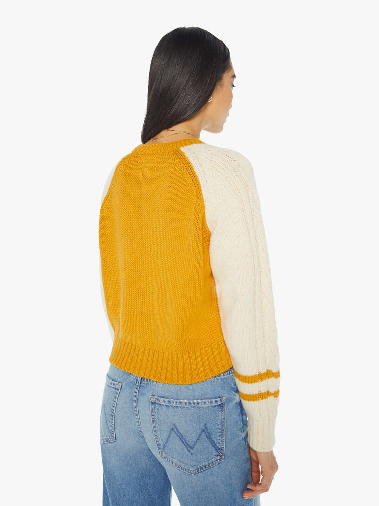 Back view of a woman letterman-inspired crewneck cardigan with raglan sleeves, ribbed hems and a slightly cropped fit in a mustard yellow with cream sleeves.