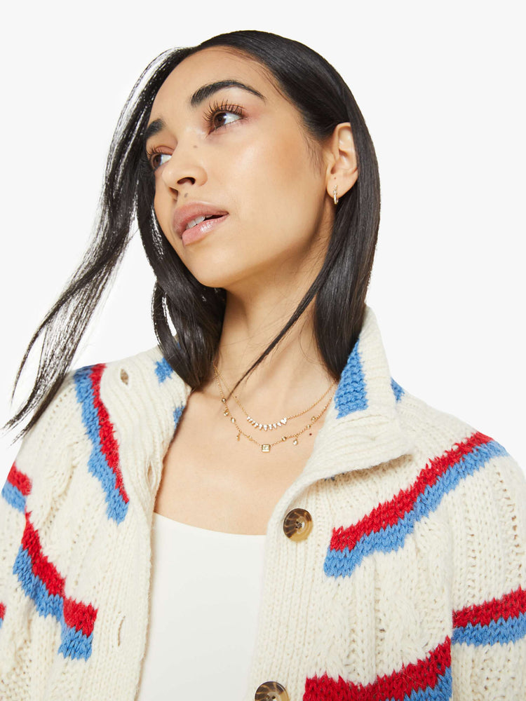 Close up view of a woman in a mock neck cardigan sweater with drop shoulders, front patch pockets, ribbed hems, buttons down the front and a slouchy fit in a cream color with red and blue stripes.