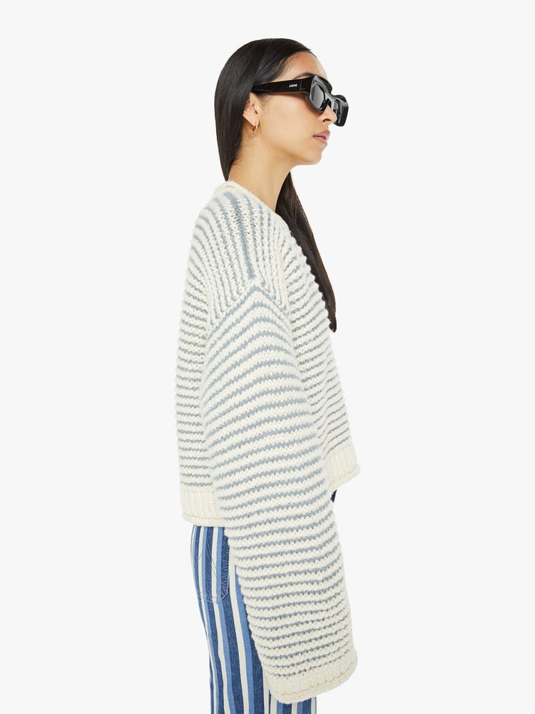 Side view of woman crewneck sweater with drop shoulders, long bell sleeves, rolled hems and a slightly cropped fit in off-white with baby blue horizontal stripes.