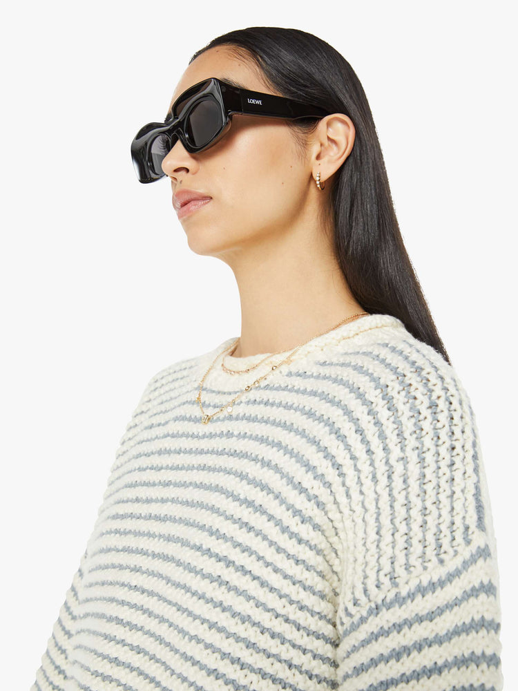 Close up view of woman crewneck sweater with drop shoulders, long bell sleeves, rolled hems and a slightly cropped fit in off-white with baby blue horizontal stripes.