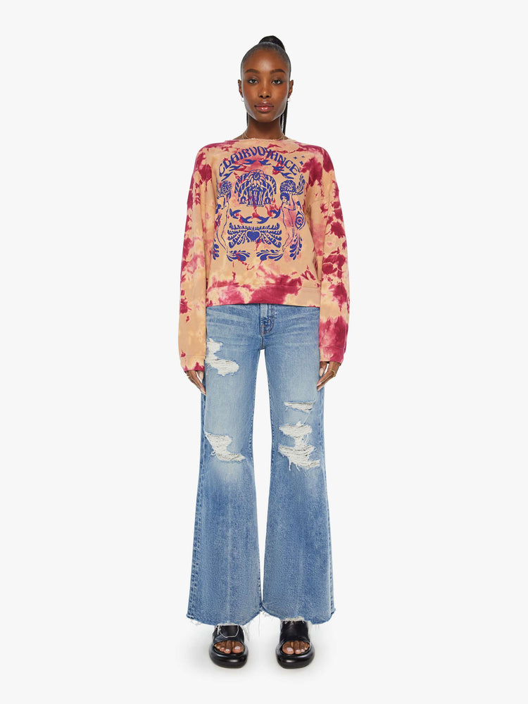 Full body view of a woman in a hand-dyed in peach and raspberry-red tie dye sweatshirt with a trippy text graphic in blue on the front.