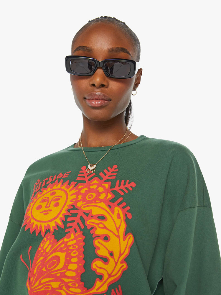 Detailed view of a woman in a green crewneck sweatshirt designed with a puff-paint graphic sun, butterfly and leaves on the front.