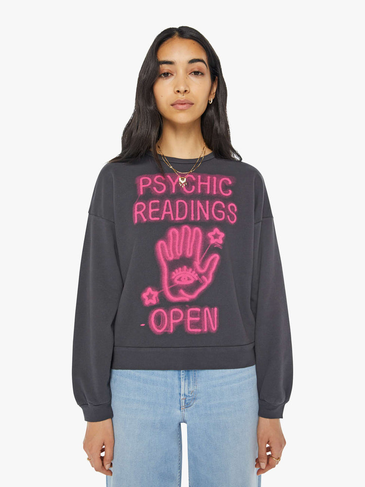 Front view of a woman in a faded black sweatshirt with a text graphic in hot pink on the front inspired by a psychic shop's neon sign.