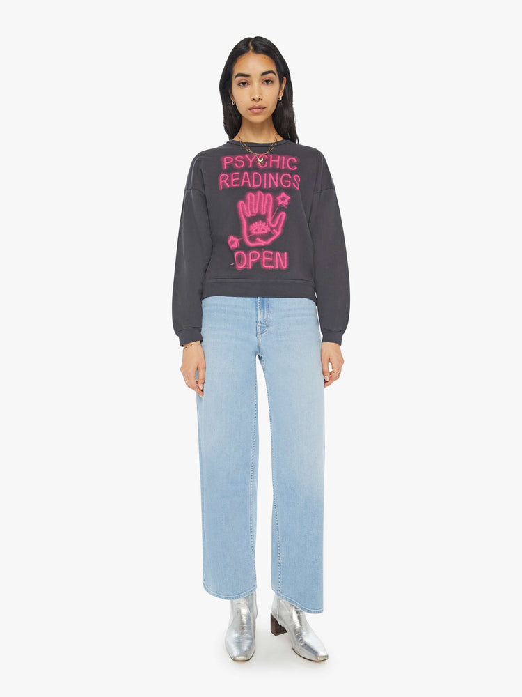 Fully body view of a woman in a faded black sweatshirt with a text graphic in hot pink on the front inspired by a psychic shop's neon sign.