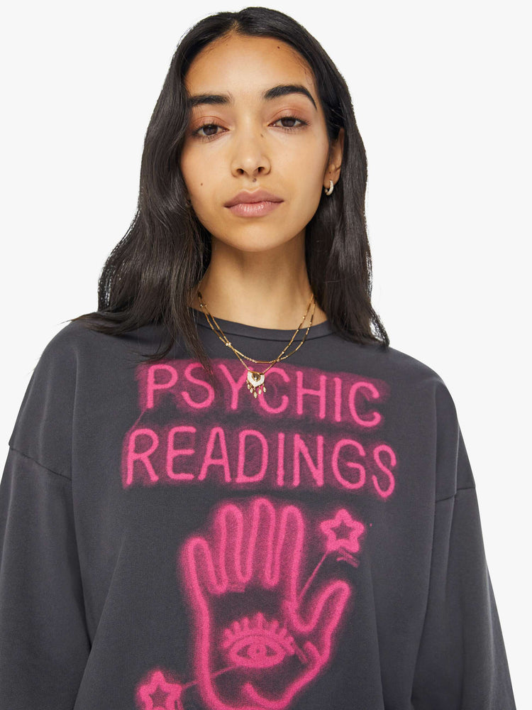 Close up view of a woman in a faded black sweatshirt with a text graphic in hot pink on the front inspired by a psychic shop's neon sign.