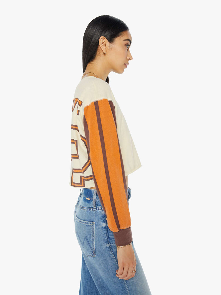 Right  side view of a woman in an athletic long sleeve tee with drop shoulders and raw hem in cream with brown sleeves and orange text graphic.