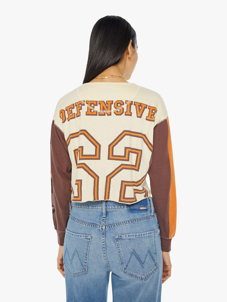 Back view of a woman in an athletic long sleeve tee with drop shoulders and raw hem in cream with brown sleeves and orange text graphic.