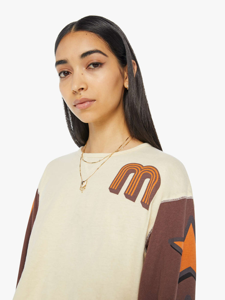 Close up view of a woman in an athletic long sleeve tee with drop shoulders and raw hem in cream with brown sleeves and orange text graphic.