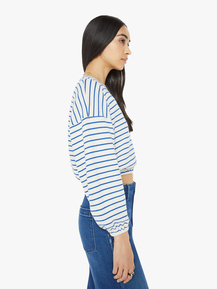 Side view of woman in a cream with blue stripes cropped long sleeve top with a crew neck, drop shoulders and elastic hems.