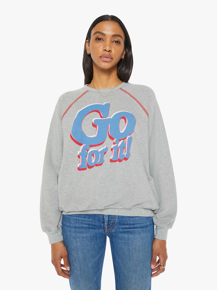 Front view of a woman in an oversized raglan sweatshirt with a crewneck, ribbed hems and a slightly cropped fit in a heathered grey with red stitching and text graphic on front.