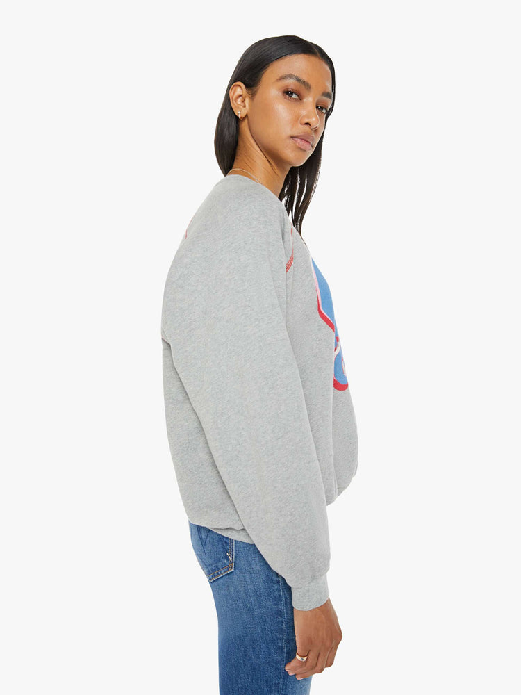 Side view of a woman in an oversized raglan sweatshirt with a crewneck, ribbed hems and a slightly cropped fit in a heathered grey with red stitching and text graphic on front.