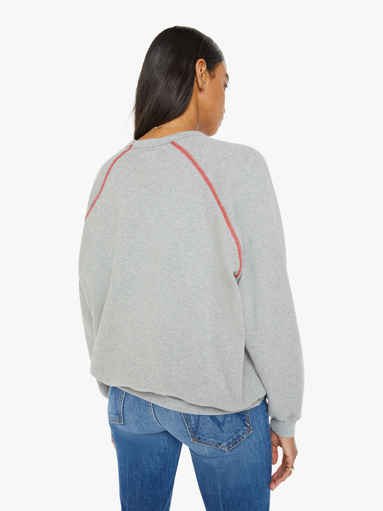 Back view of a woman in an oversized raglan sweatshirt with a crewneck, ribbed hems and a slightly cropped fit in a heathered grey with red stitching and text graphic on front.