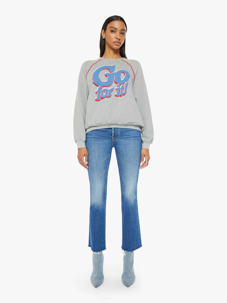 Full body view of a woman in an oversized raglan sweatshirt with a crewneck, ribbed hems and a slightly cropped fit in a heathered grey with red stitching and text graphic on front.