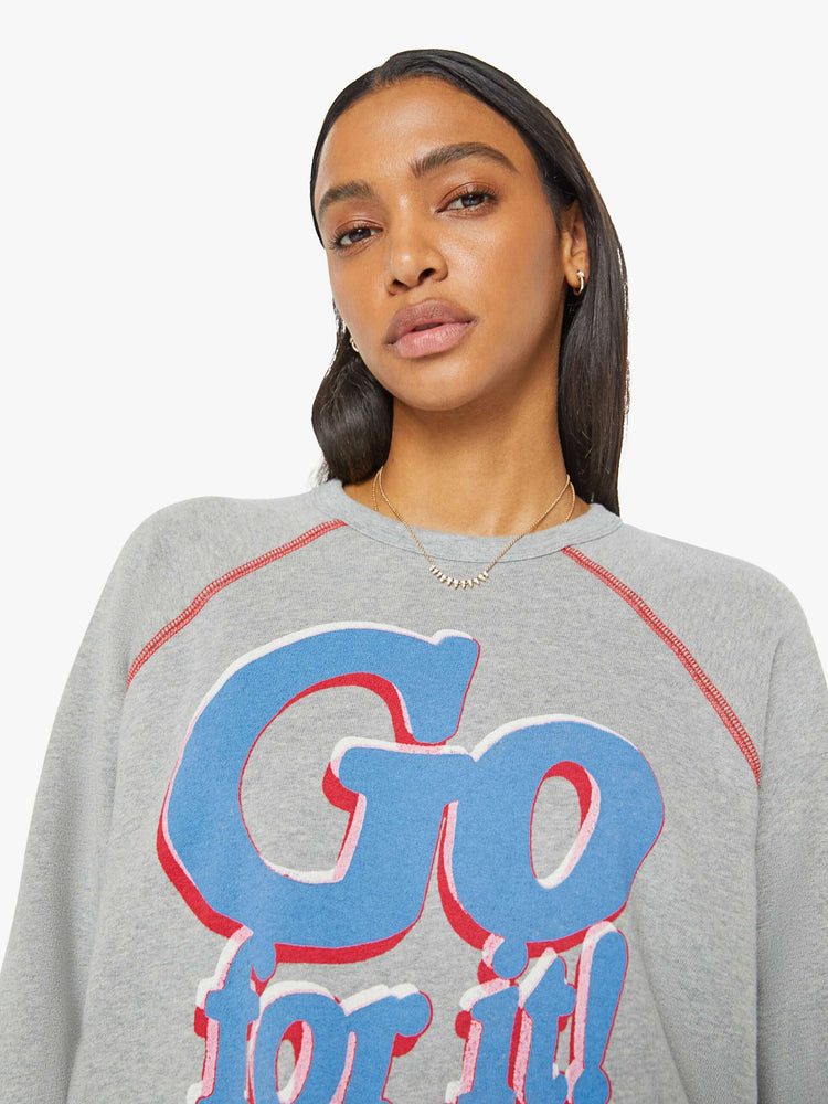 Close up view of a woman in an oversized raglan sweatshirt with a crewneck, ribbed hems and a slightly cropped fit in a heathered grey with red stitching and text graphic on front.