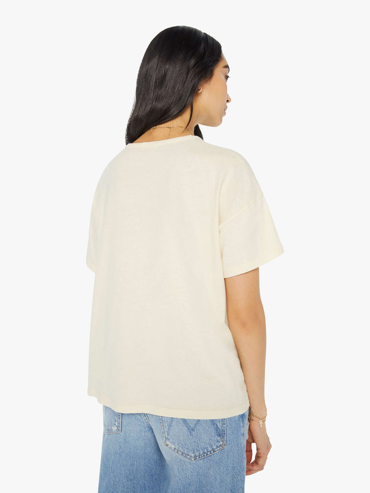 Back view of a woman in a oversized tee with drop shoulders and a loose, boxy fit in a cream color with a faded text graphic in purple.