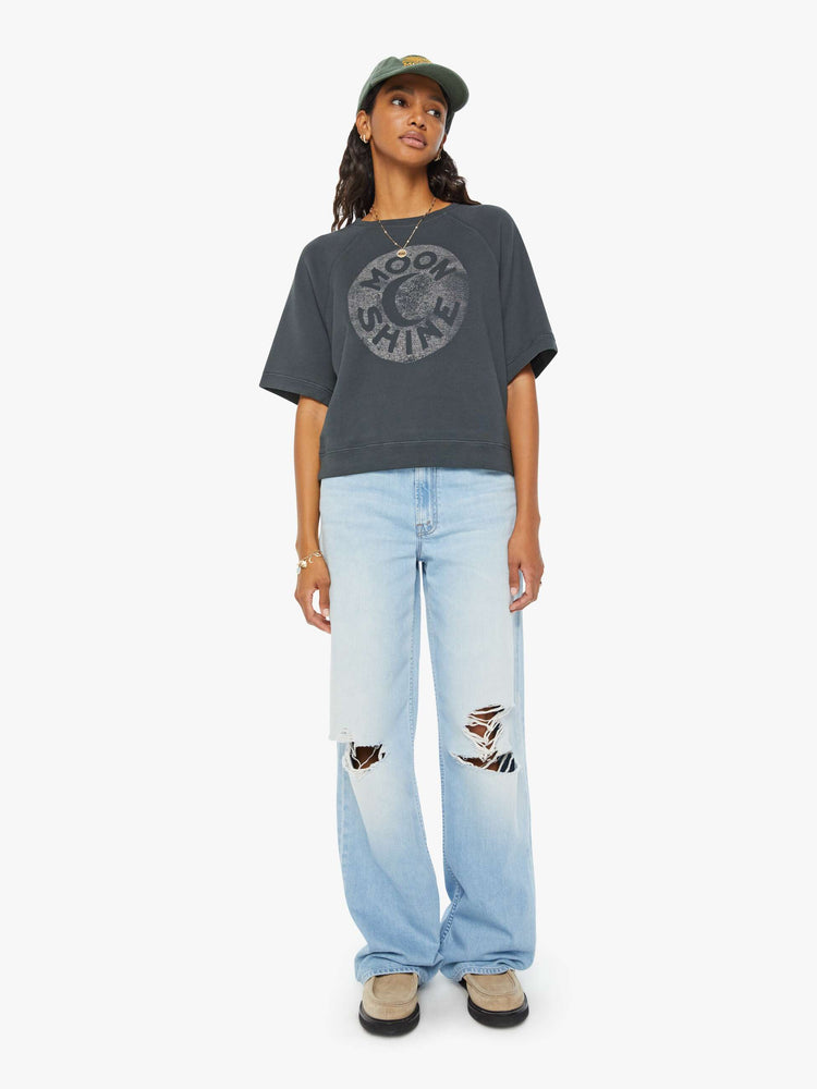 Full body view of a woman dark grey short sleeve raglan sweatshirt with ribbed hems and a loose, boxy fit with a faded moon graphic with text in white on the front.