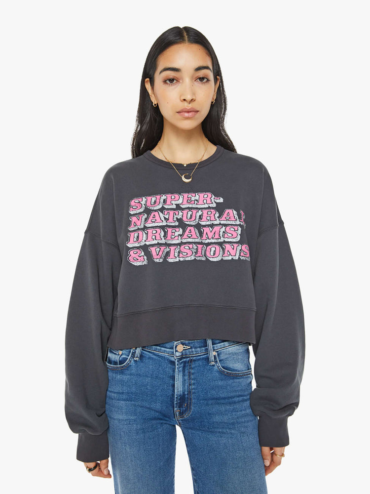 Front view of a woman in a faded black  sweatshirt with drop shoulders, super long sleeves, ribbed hems and a wide, cropped fit with pink text on front.