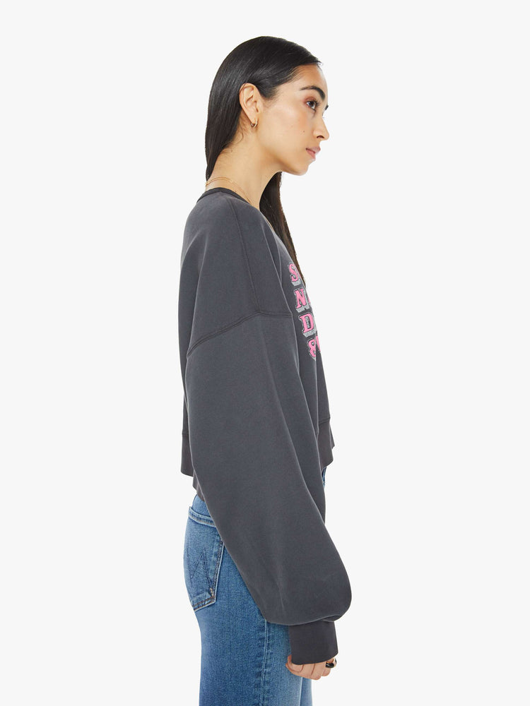 Side view of a woman in a faded black  sweatshirt with drop shoulders, super long sleeves, ribbed hems and a wide, cropped fit with pink text on front.