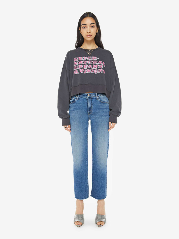 Full body view of a woman in a faded black  sweatshirt with drop shoulders, super long sleeves, ribbed hems and a wide, cropped fit with pink text on front.