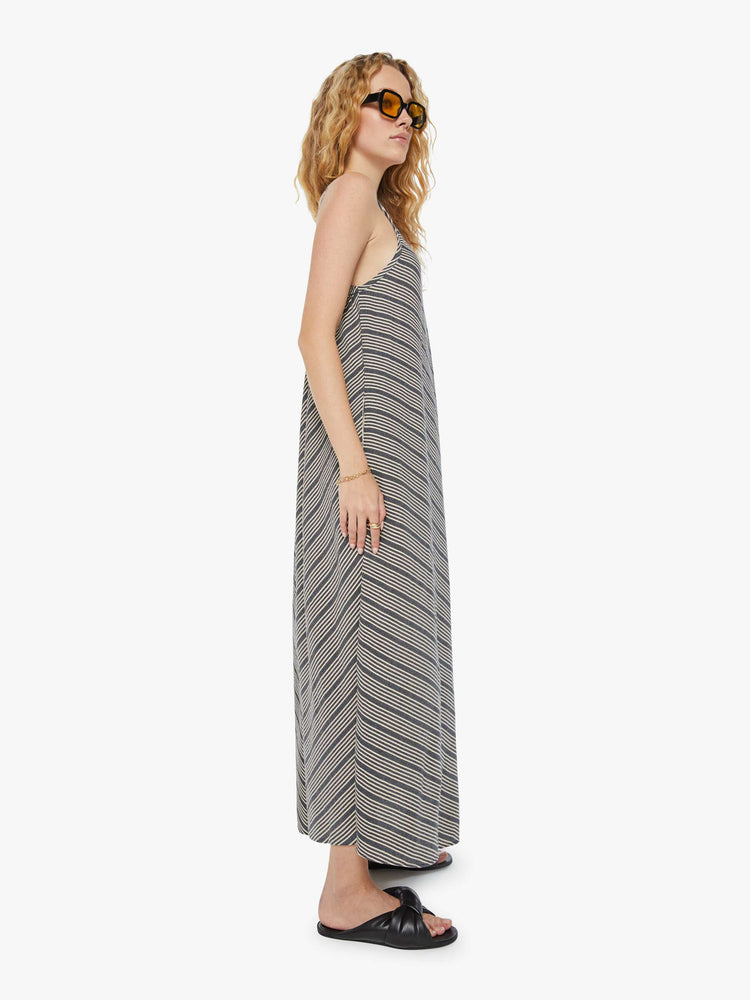 Side view of a woman in a grey and white striped  maxi dress with a V-neck, thin spaghetti straps that tie in the back, an ankle-length hem and a flowy fit. 