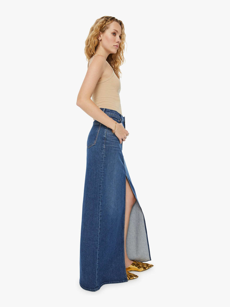 Side view of a woman in a dark blue denim maxi skirt with a high rise, thigh-high front slit and a floor-length hem.