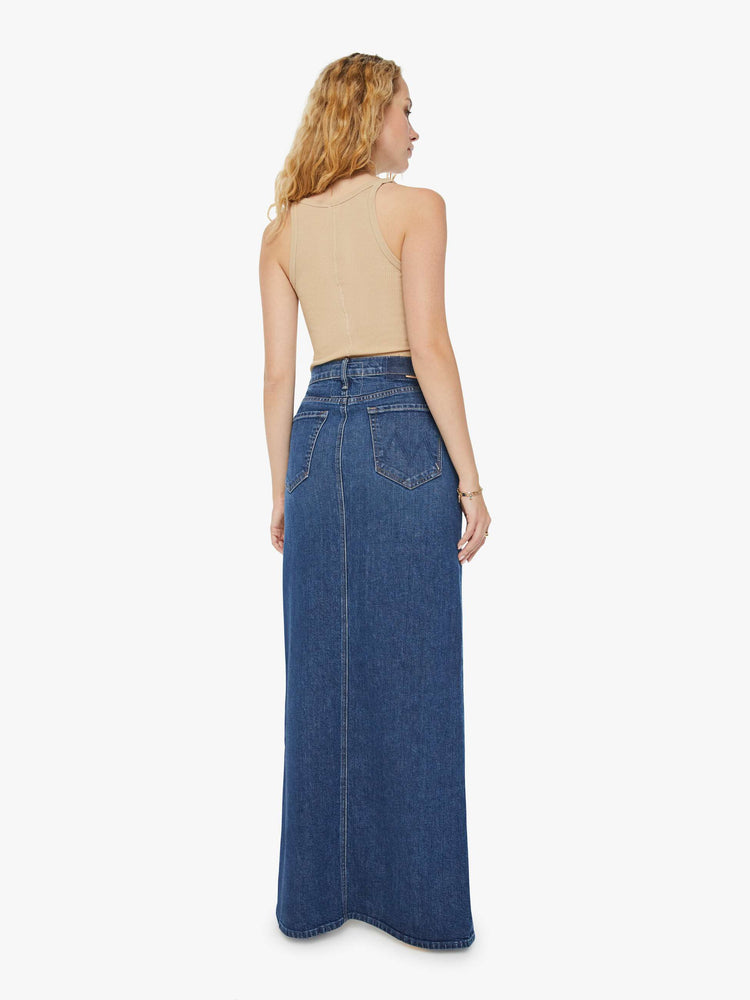 Back view of a woman in a dark blue denim maxi skirt with a high rise, thigh-high front slit and a floor-length hem.