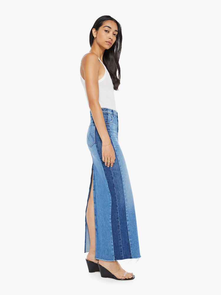 Side view of a woman in a denim maxi skirt with a high rise, thigh-high back slit and a frayed ankle-length hem in a mid blue and dark panels down the outer leg.