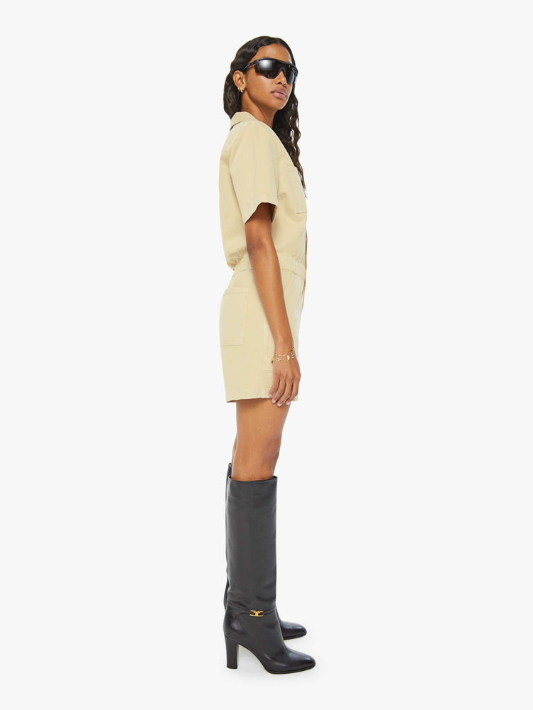 Side view of a woman in a light khaki  V-neck romper with a notched collar, short sleeves, patch pockets, buttons down the front and an elastic waistband. 